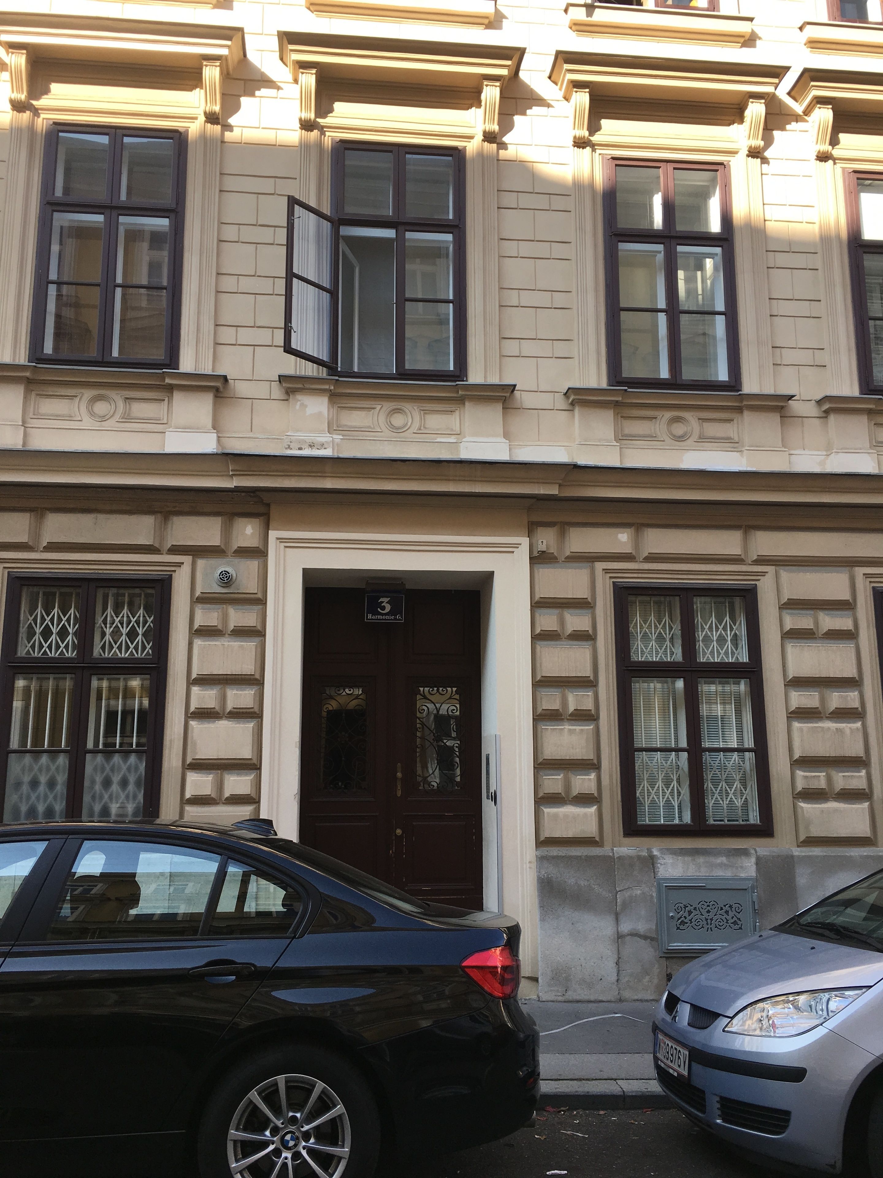 A building with a classic architectural style, featuring a central entrance with a dark wooden door and a small balcony above it. There are two cars parked in front of the building.