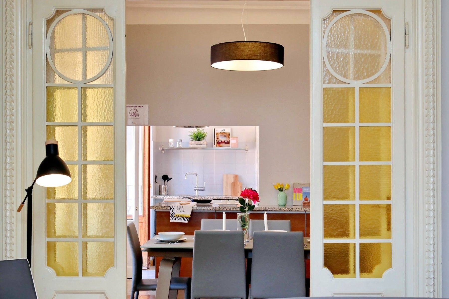 A dining area with a table set for two, a hanging light fixture, and a view into a kitchen with a white countertop and various items on the shelves.