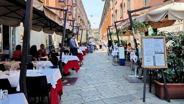 An outdoor dining area with tables set up along a cobblestone street, surrounded by buildings with balconies. - JOIVY Austere Elegance (Milan)