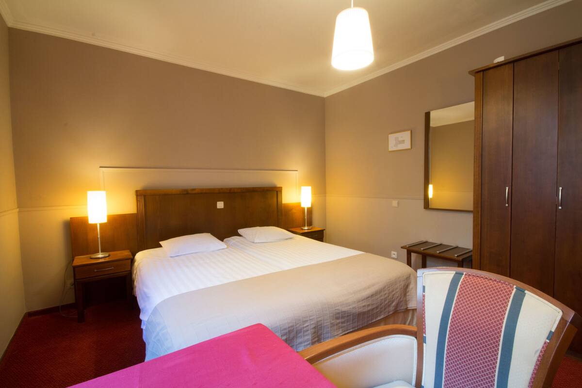 A hotel room with a large bed, two bedside lamps, a wooden wardrobe, and a chair with a striped cushion.
