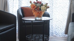 A round table with a glass vase containing flowers, positioned between two black armchairs with a patterned cushion, in a room with patterned curtains and wooden flooring. - My Way (La Spezia)