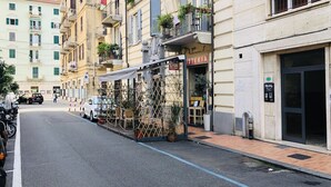 A street with a row of buildings, including a restaurant named "OSTERIA" with outdoor seating and potted plants. - My Way (La Spezia)