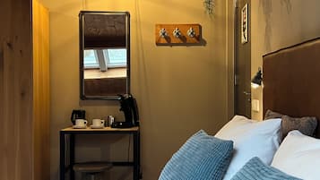Habitación de hotel con una cama, un escritorio con cafetera, una silla, un taburete pequeño, una ventana y un estante de pared con tres objetos decorativos.