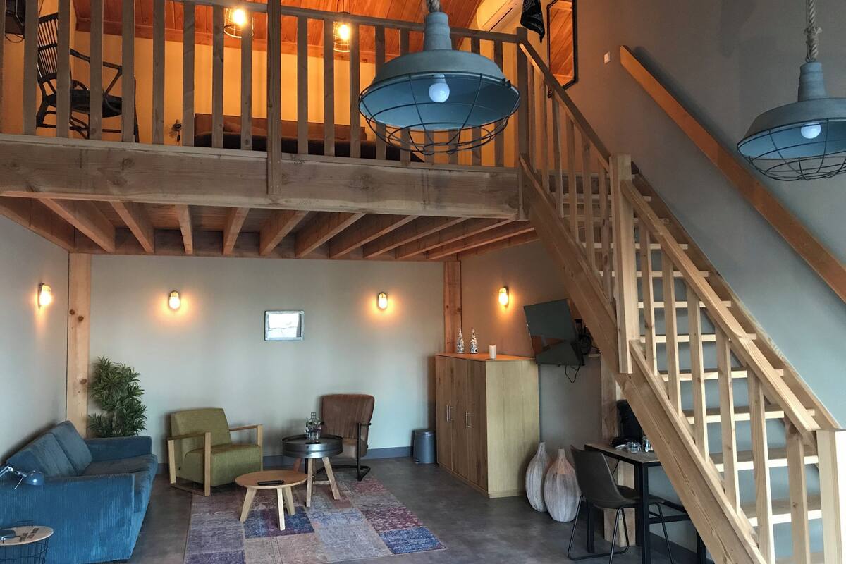 A spacious loft with a high ceiling, exposed wooden beams, a staircase with a wooden railing, and a cozy seating area with a sofa, chairs, and a small table.