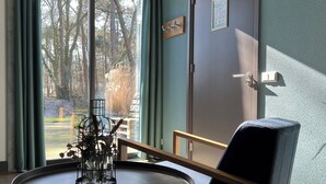 Une pièce avec une grande fenêtre, une chaise, une petite table sur laquelle se trouve un vase, et une porte ornée d’un cadre de photo.