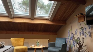 Une pièce chaleureuse avec un plafond en pente, des poutres en bois, une grande fenêtre, un fauteuil jaune, un fauteuil bleu, une petite table avec des tasses et une plante en pot.