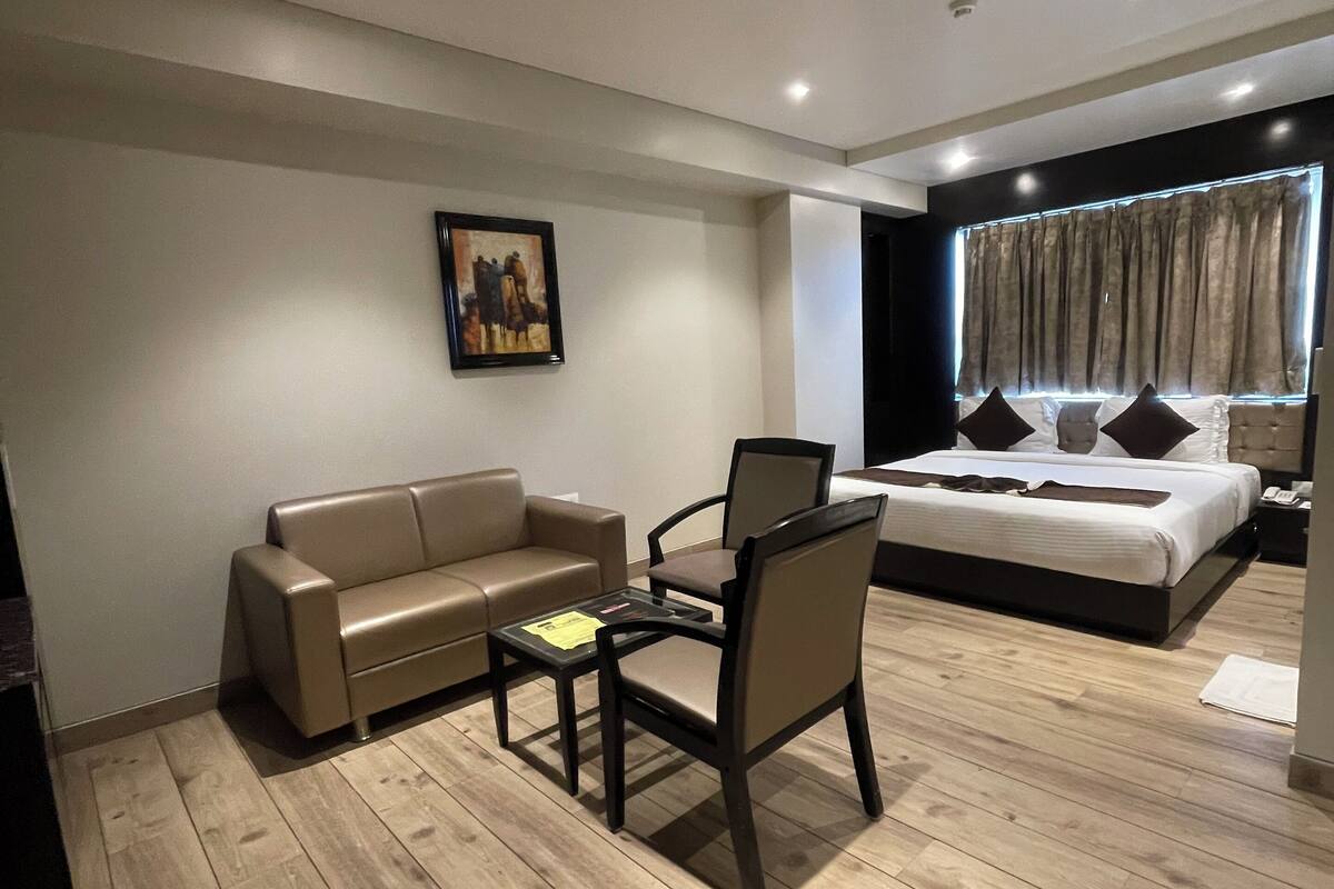 A modern hotel room with a wooden floor, a sofa, a chair, a small table, a bed with a headboard, a painting on the wall, and a window with curtains.