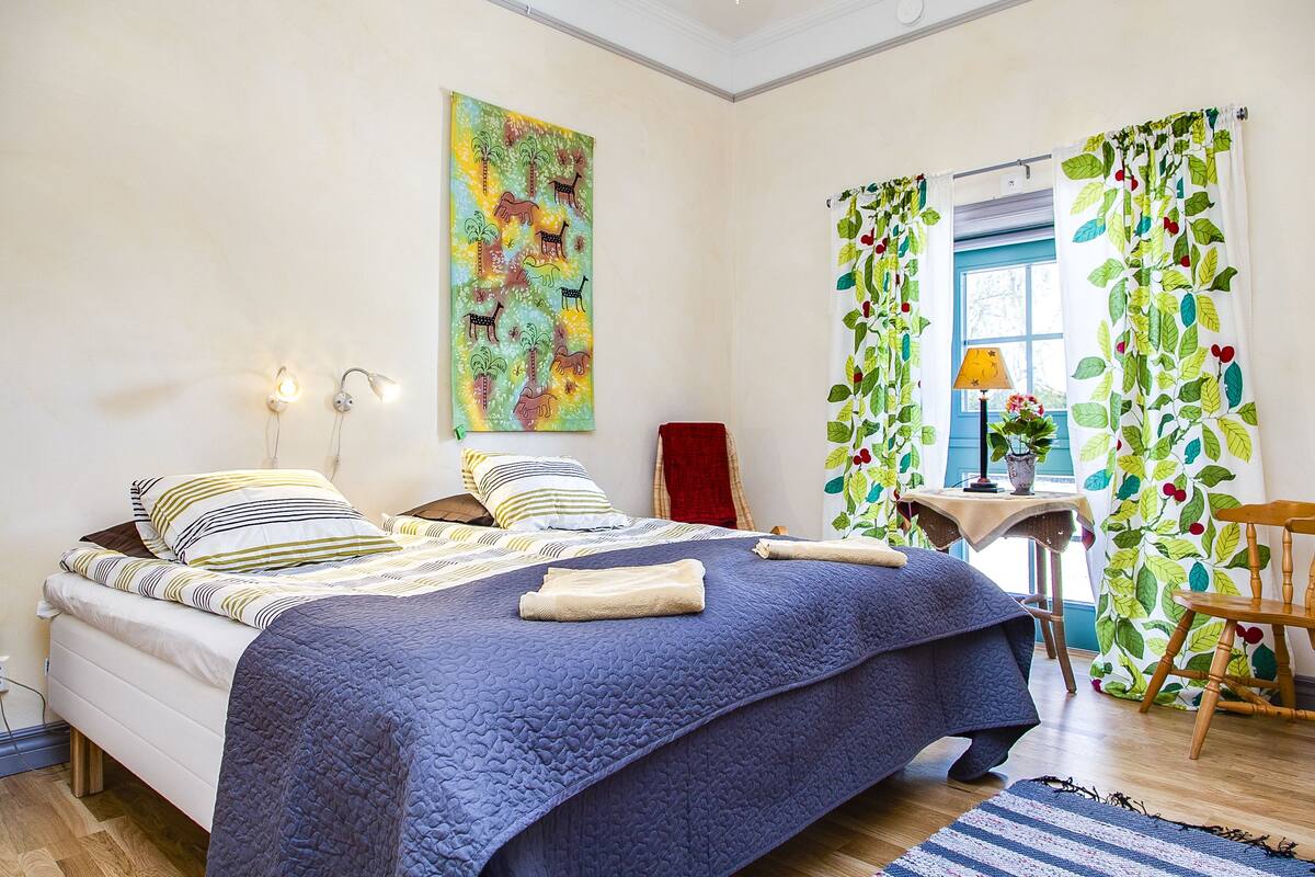 A bedroom with a bed, a bedside table with a lamp, a chair, and a window with curtains.