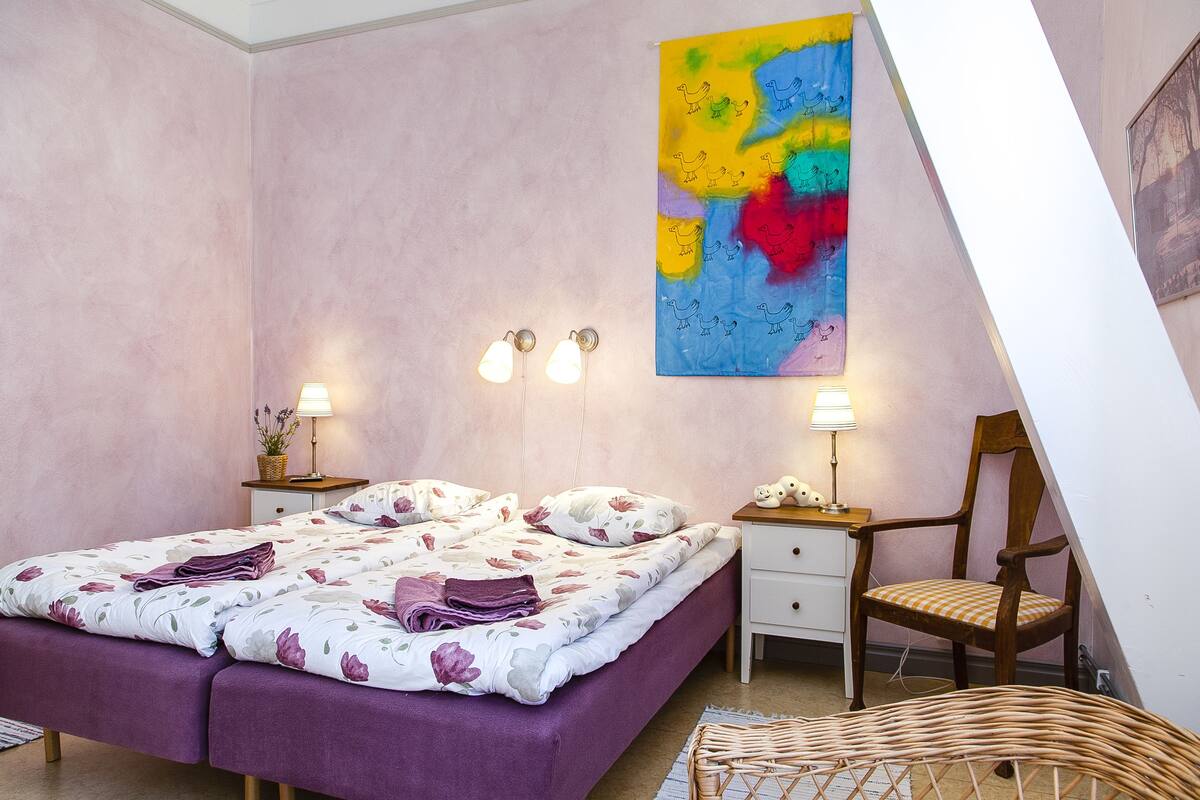 A bedroom with a bed, two bedside tables, a chair, a lamp, and a colorful abstract painting on the wall.