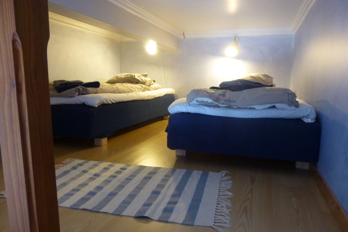 A small, dimly lit room with two single beds, a striped rug, and a wooden floor.