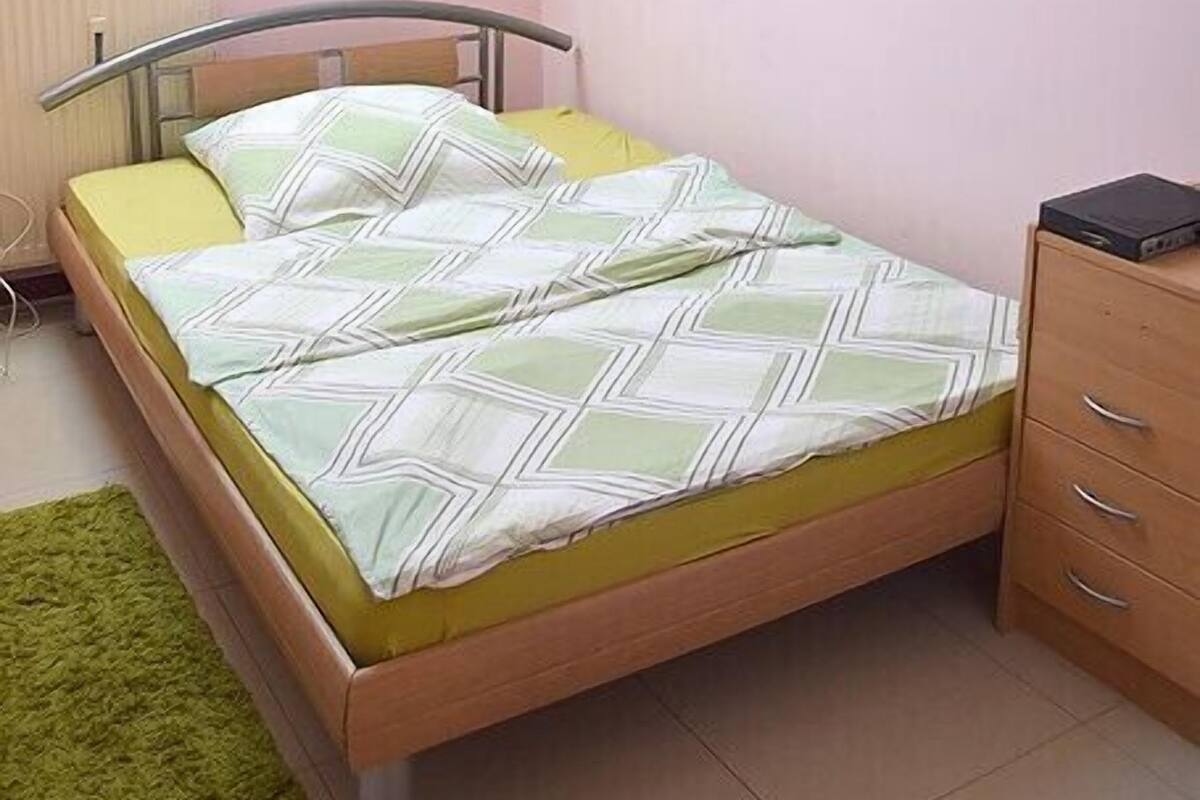 A single-bed room with a sloped ceiling, a radiator, a green rug, a wooden nightstand, and a painting of tulips on the wall.