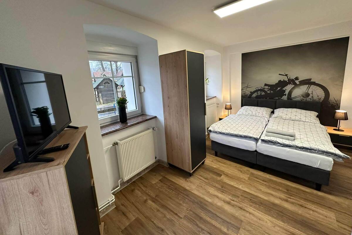 A modern bedroom with a bed, bedside tables, a TV, a radiator, and a window with a view of houses.