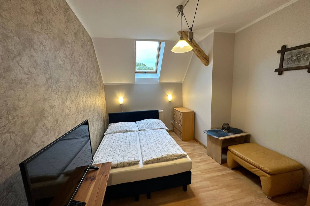 A bedroom with a bed, bedside table, bench, and a desk with a computer. There is a window and a ceiling fan.