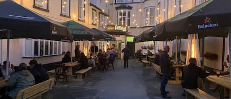 An outdoor seating area with tables and benches, surrounded by buildings with string lights.