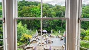 A view from a window of a deck with wooden tables and chairs, surrounded by lush greenery and trees. - The Hand Hotel Llangollen by Compass Hospitality (Llangollen)