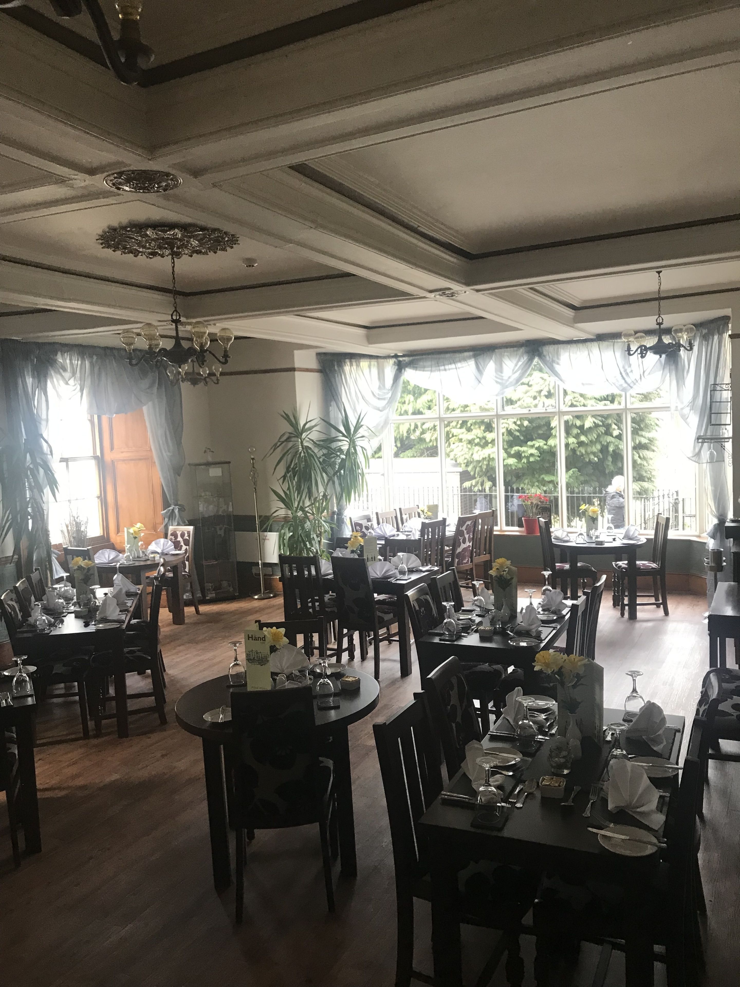 Un comedor con varias mesas y sillas, ventanas amplias con cortinas y un candelabro.