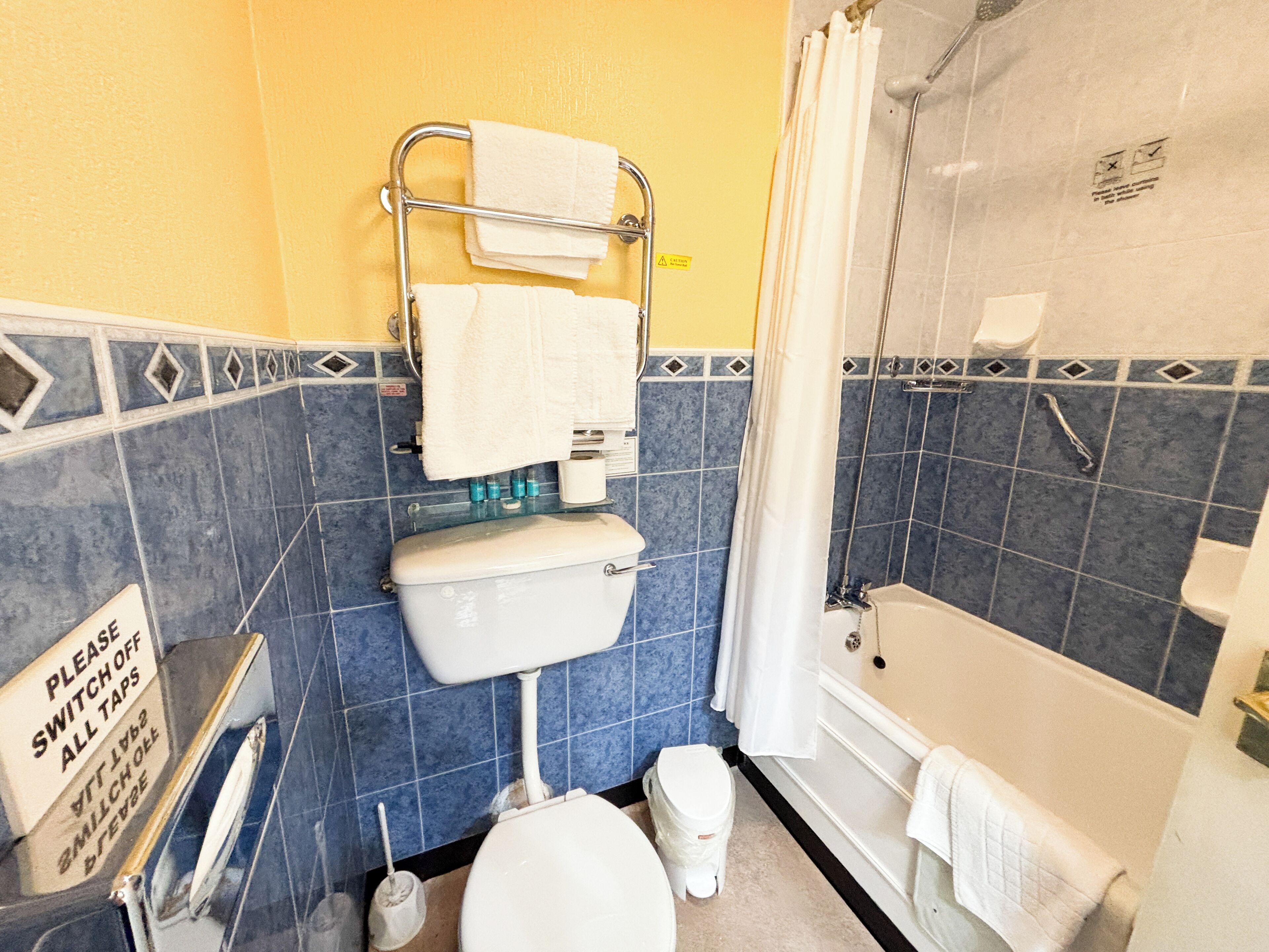 A bathroom with a toilet, a shower, and a bathtub. There is a sign on the wall that says "Please switch off all taps."