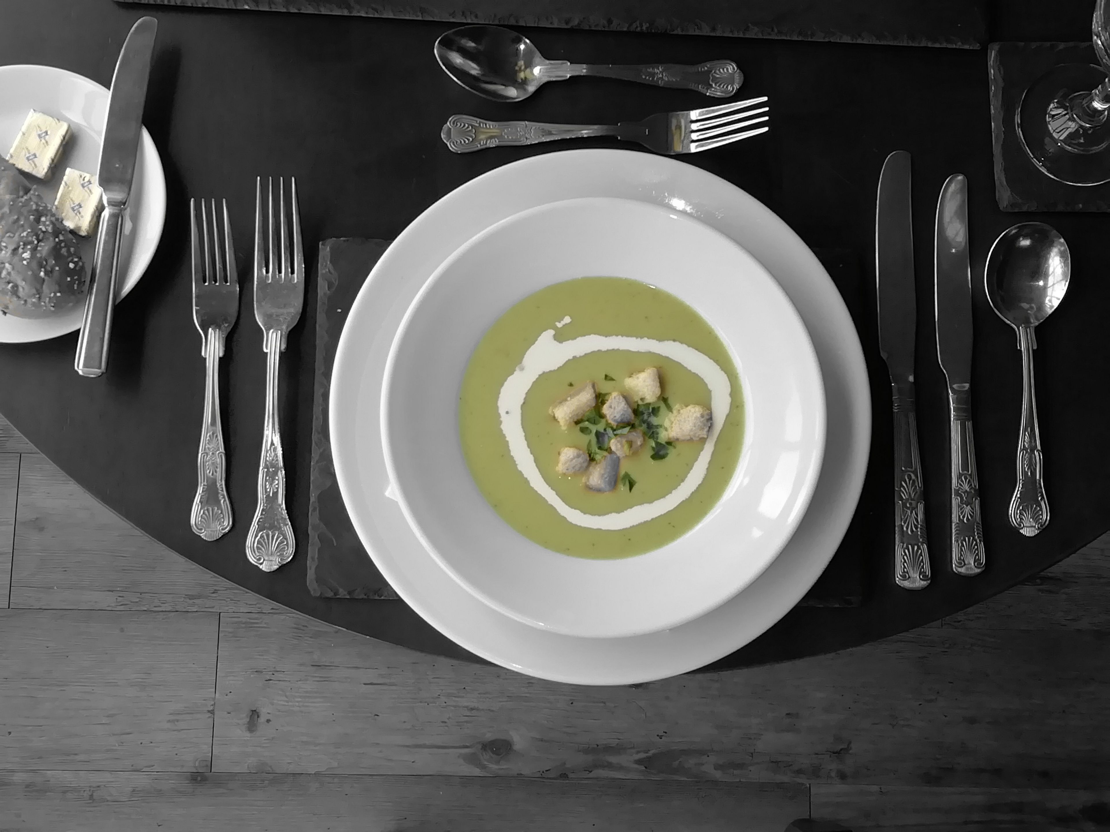 Une table à manger avec une assiette blanche contenant une soupe verte, garnie de croûtons et d’herbes, entourée de couverts en argent.