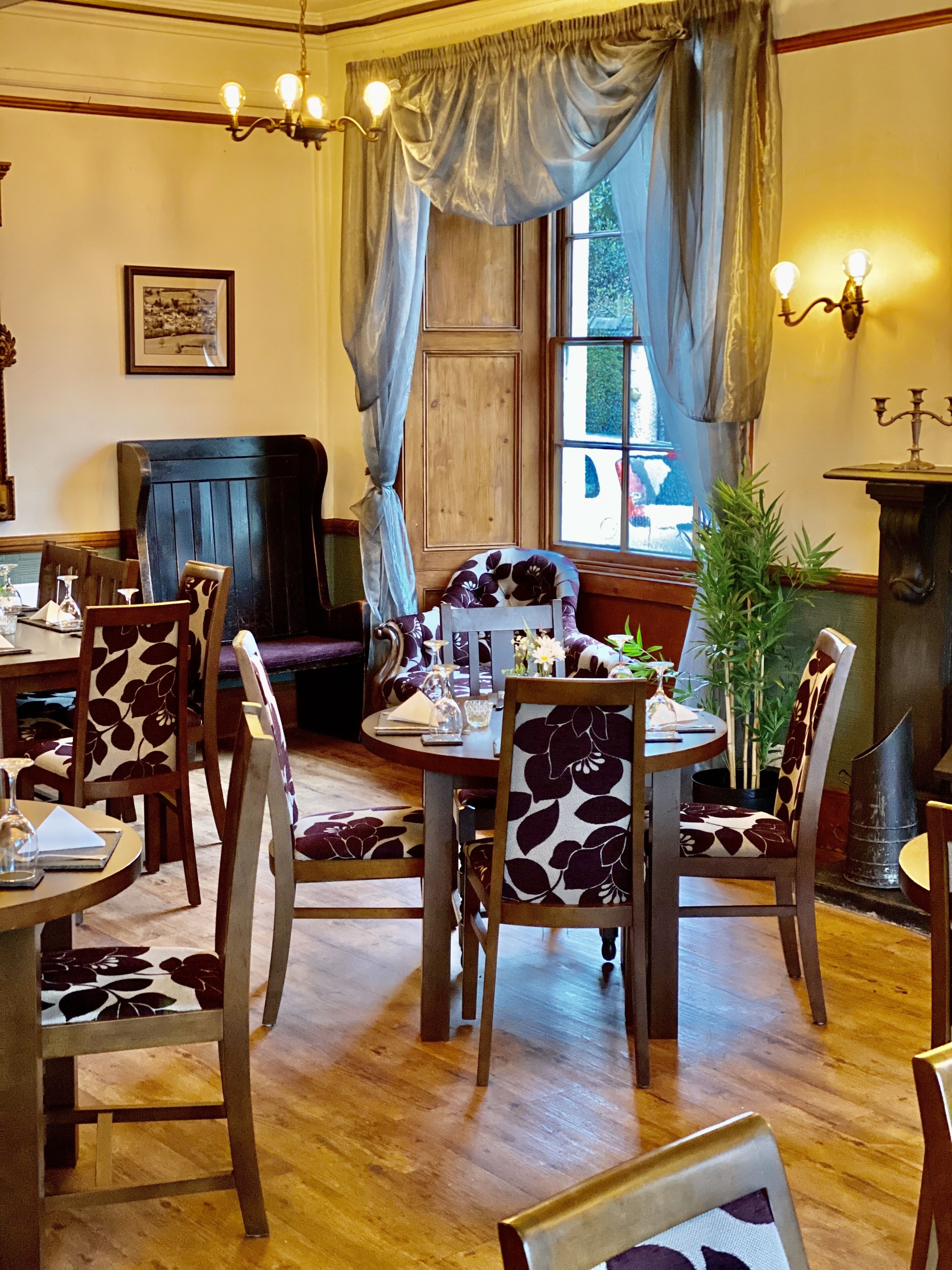 Un comedor con mesas y sillas de madera, una araña y una ventana con cortinas.