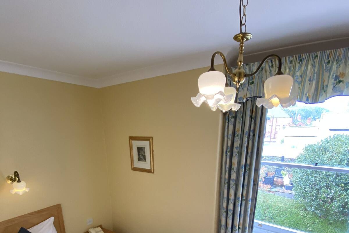 A hotel room with a large bed, a bedside table with a phone, a hanging light fixture, and a window with curtains.