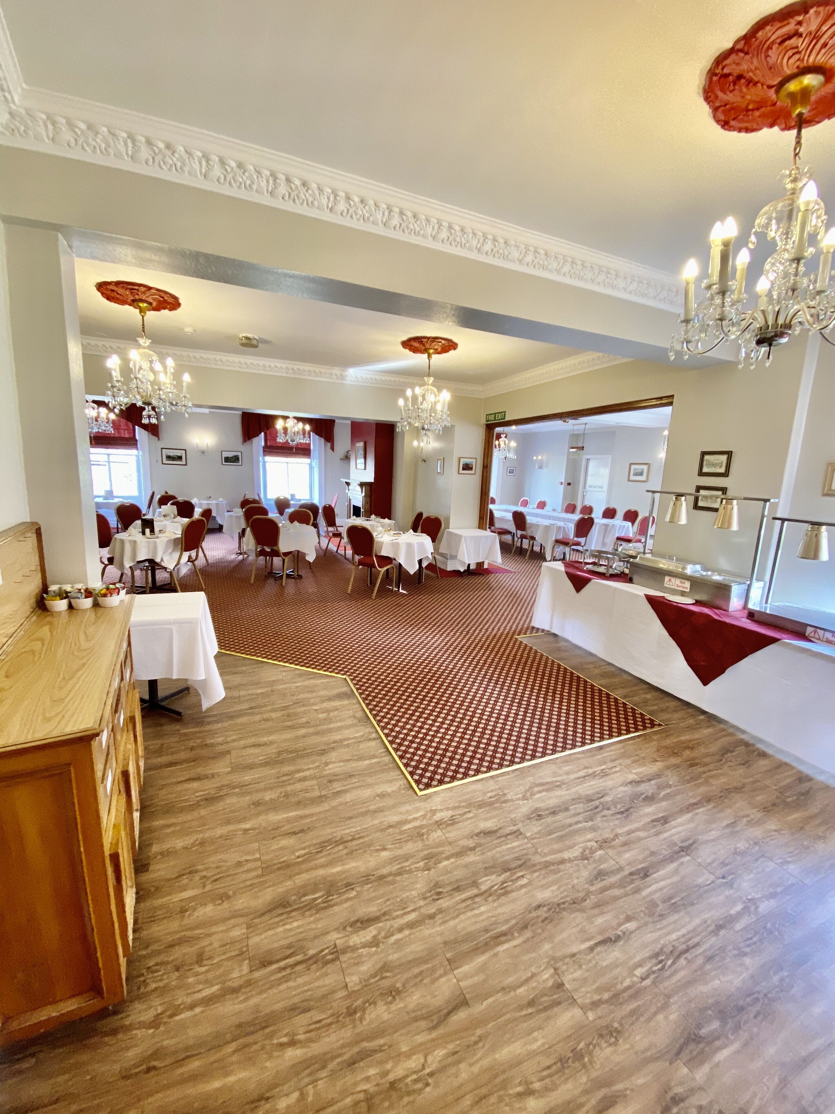 A spacious banquet hall with multiple round tables and chairs, a patterned carpet, and elegant chandeliers.
