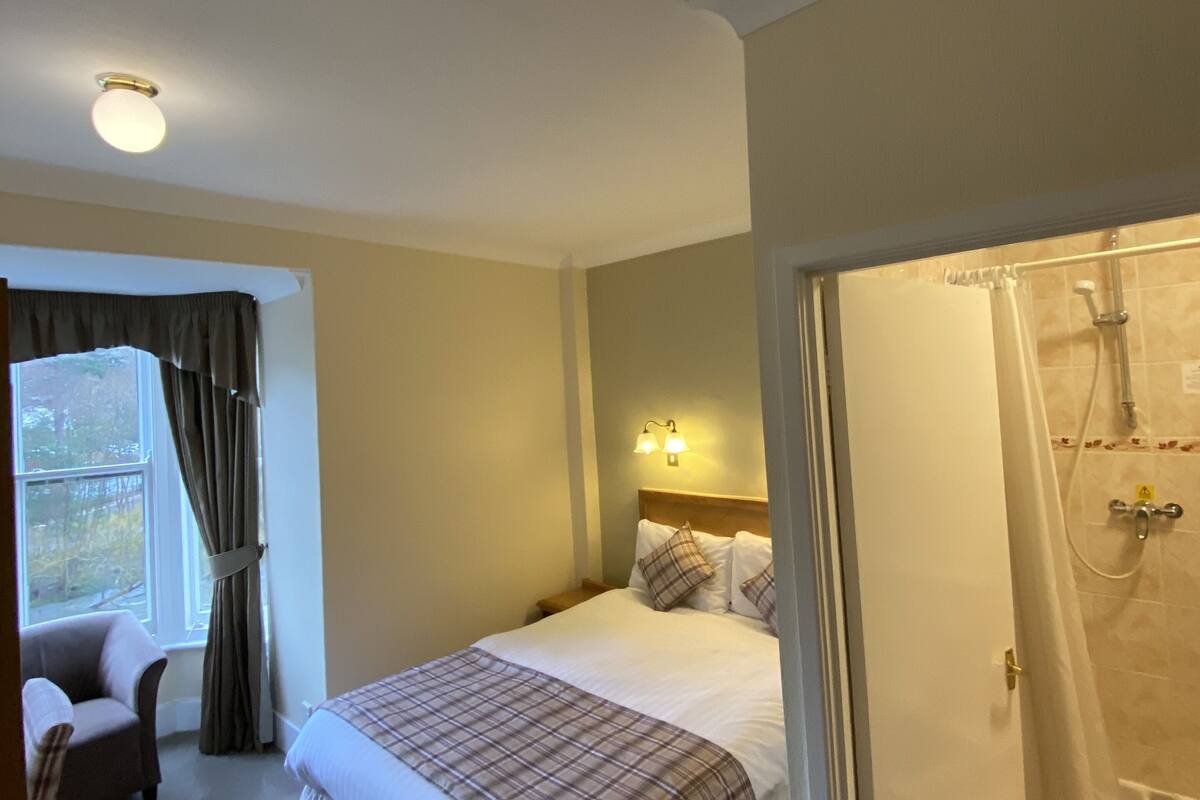 A hotel room with a bed, a chair, a window with curtains, a shower area, and a wall-mounted light fixture.