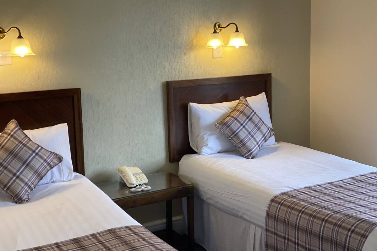 A hotel room with two beds, each with a wooden headboard, a bedside table with a phone, and two wall-mounted light fixtures.