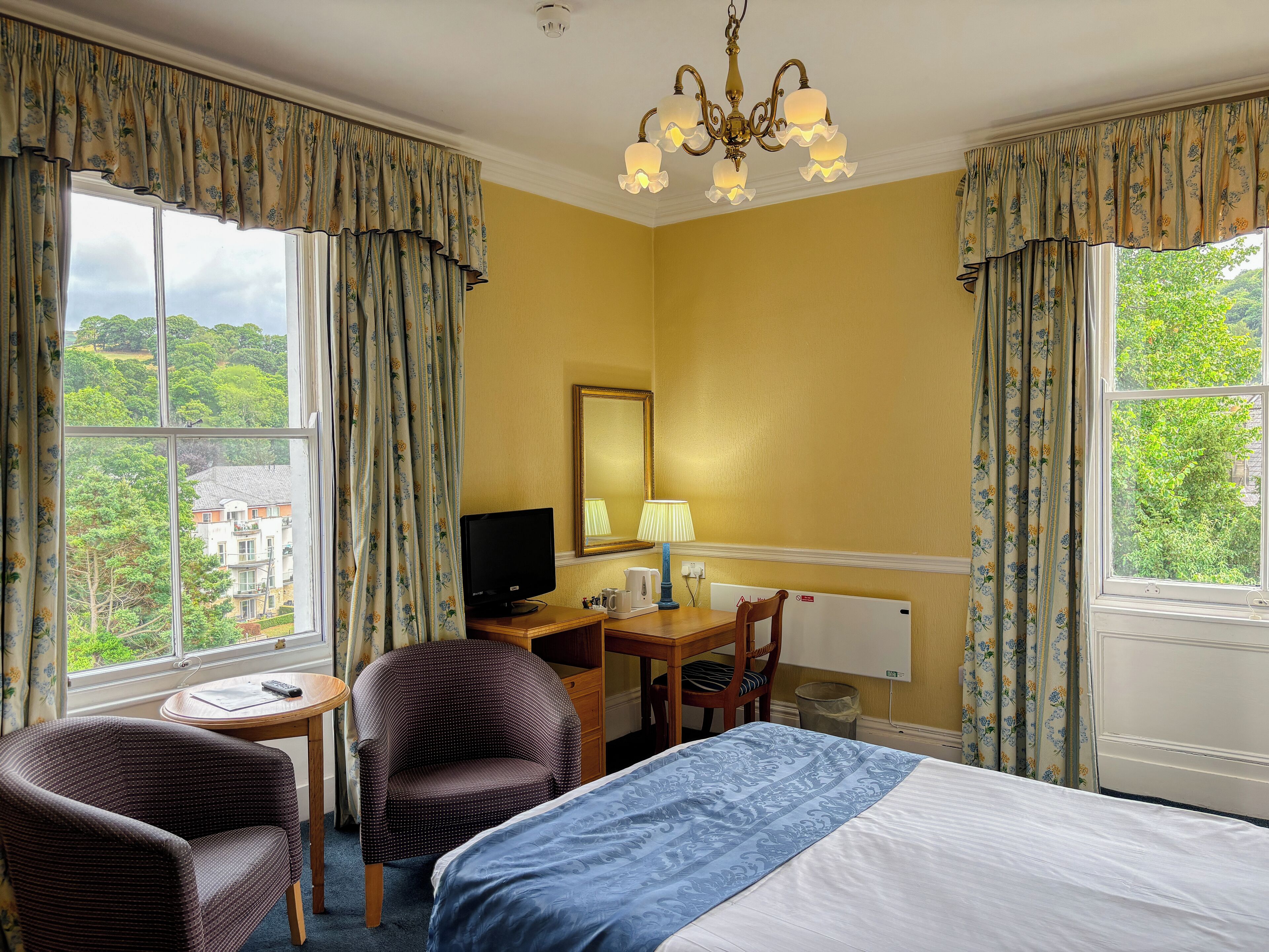 A hotel room with a bed, a desk with a computer, a chair, and a view of greenery outside.