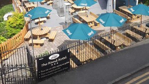 Un espace extérieur aménagé avec plusieurs tables et chaises, situé au bord de l’eau, ombragé par des parasols bleus, et sur lequel on peut voir une enseigne indiquant « The Land Hotel ».