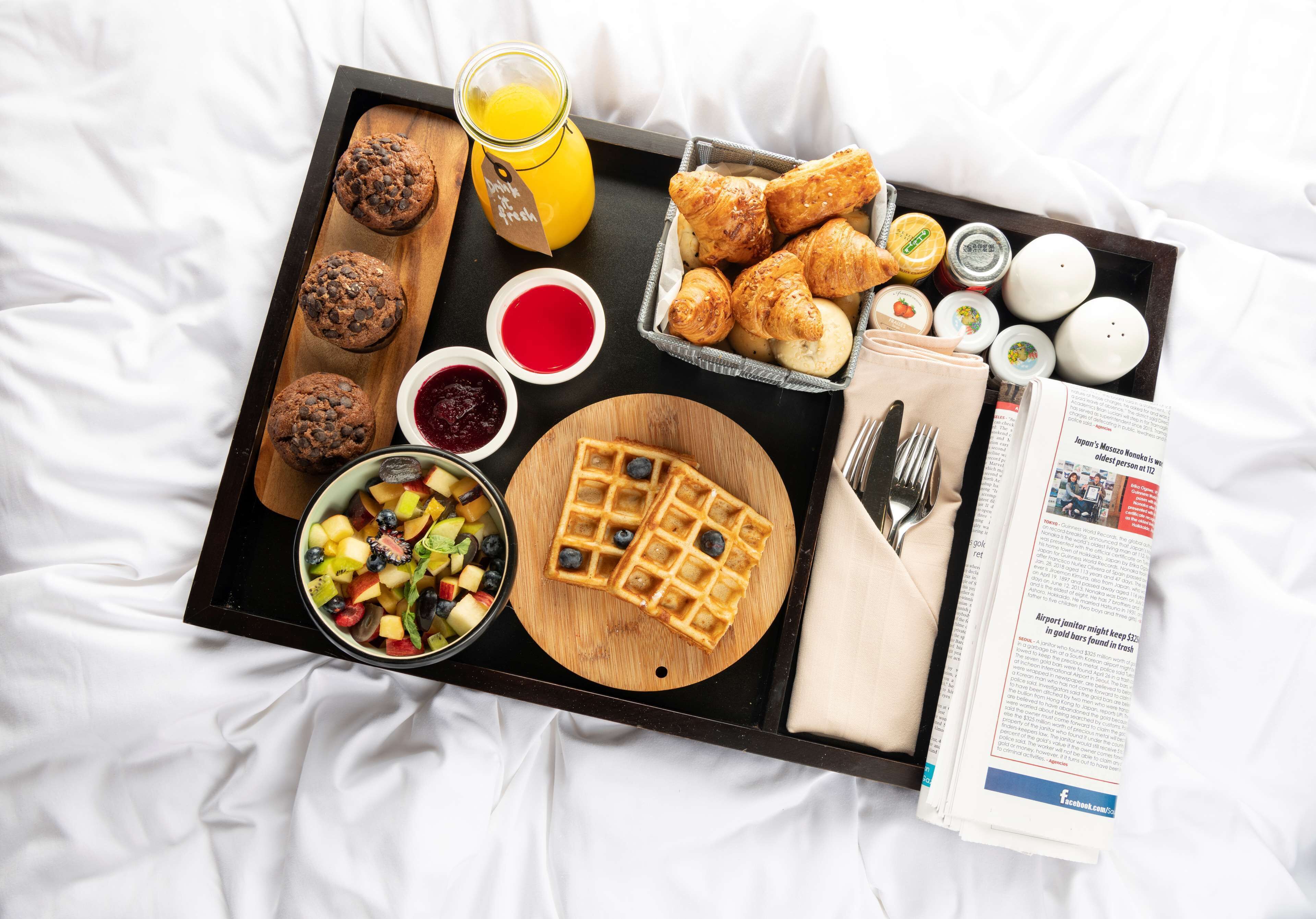 A breakfast tray with croissants, waffles, muffins, and a fruit bowl on a bed with a newspaper.
