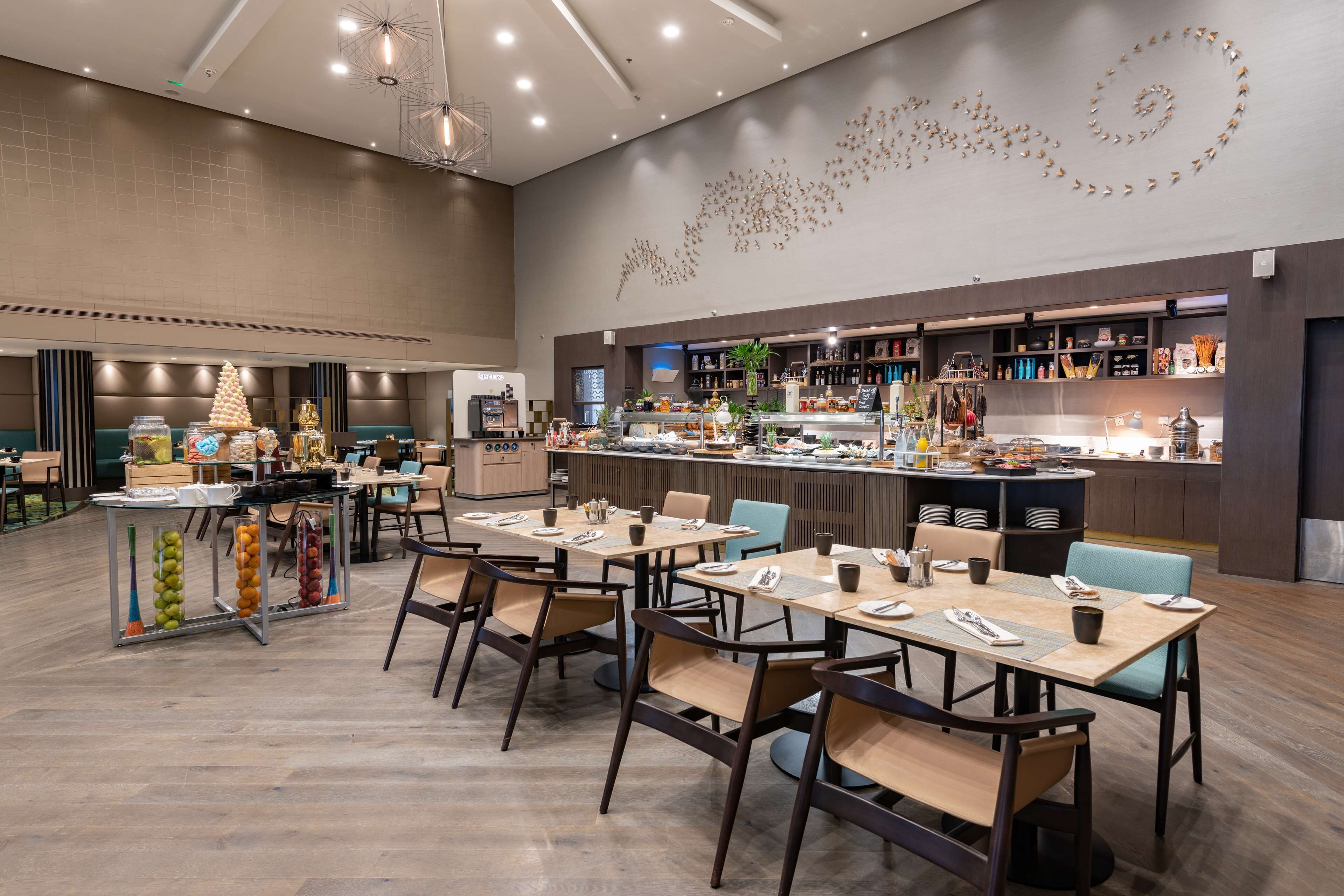 A modern restaurant with multiple dining tables and chairs, a kitchen area, and a decorative wall.