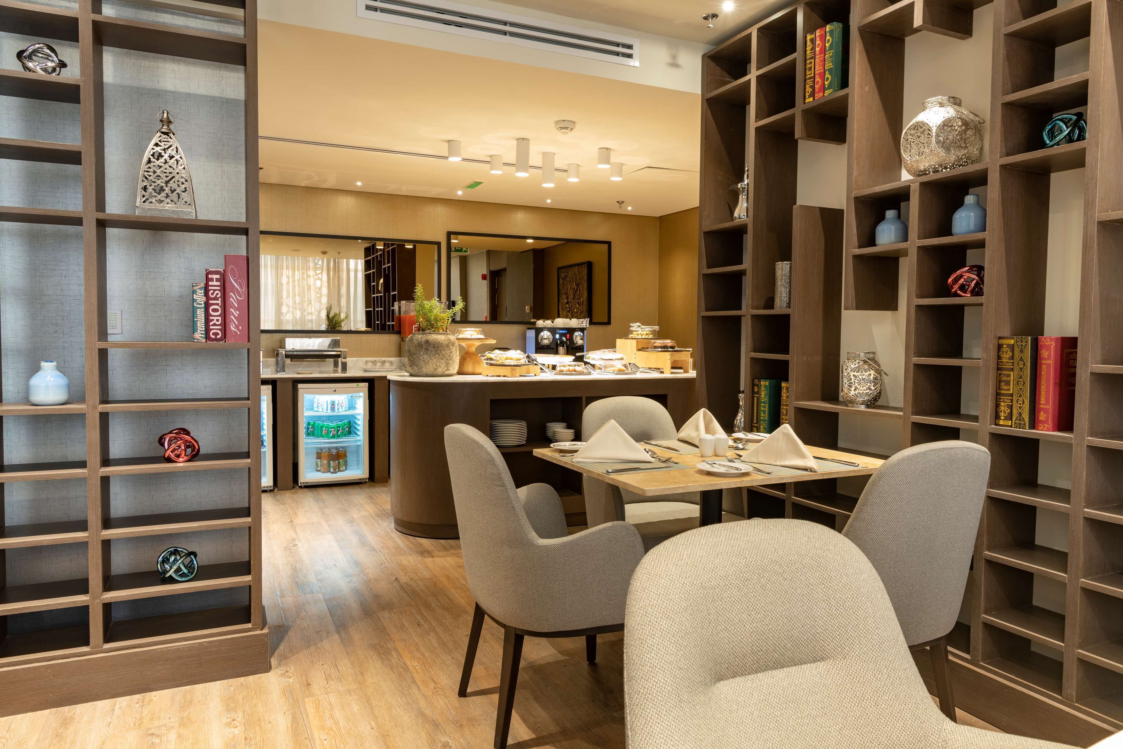 A modern hotel dining area with a wooden bookshelf, a dining table with chairs, and a refrigerator in the background.