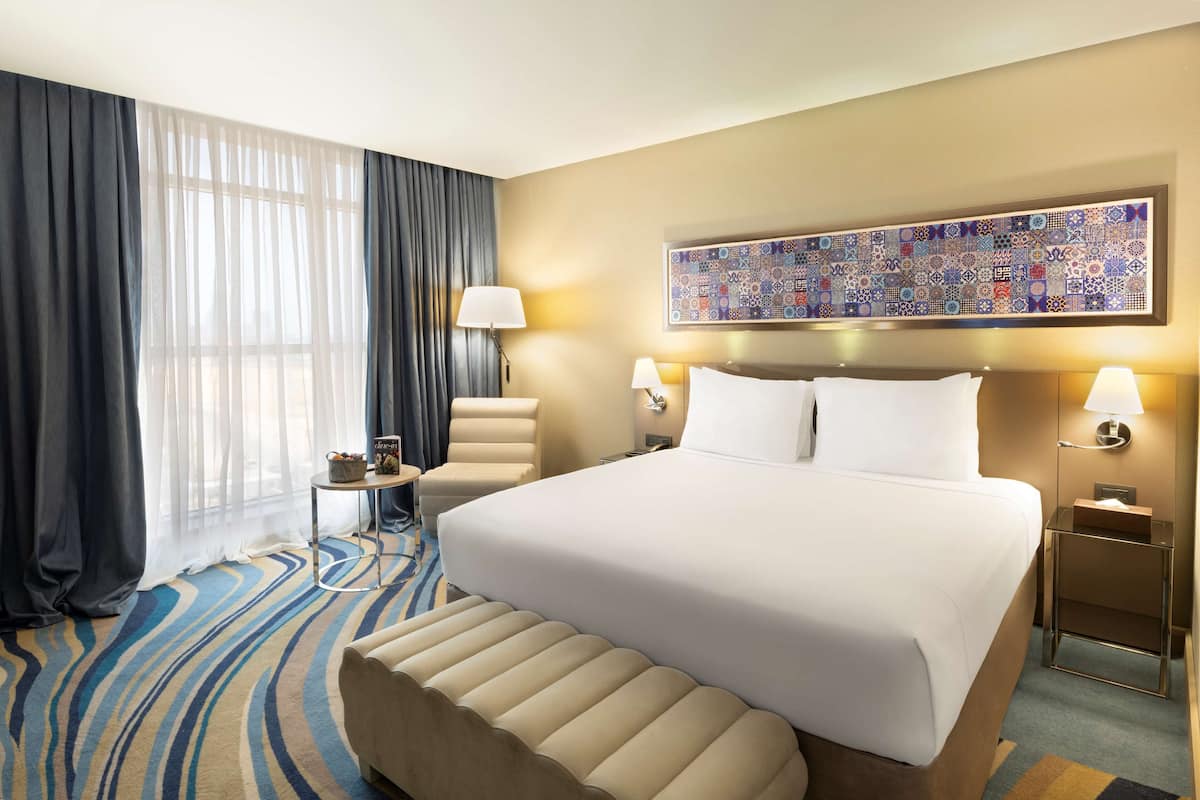 A hotel room with a large bed, a desk, a chair, a window with curtains, and a decorative wall piece.