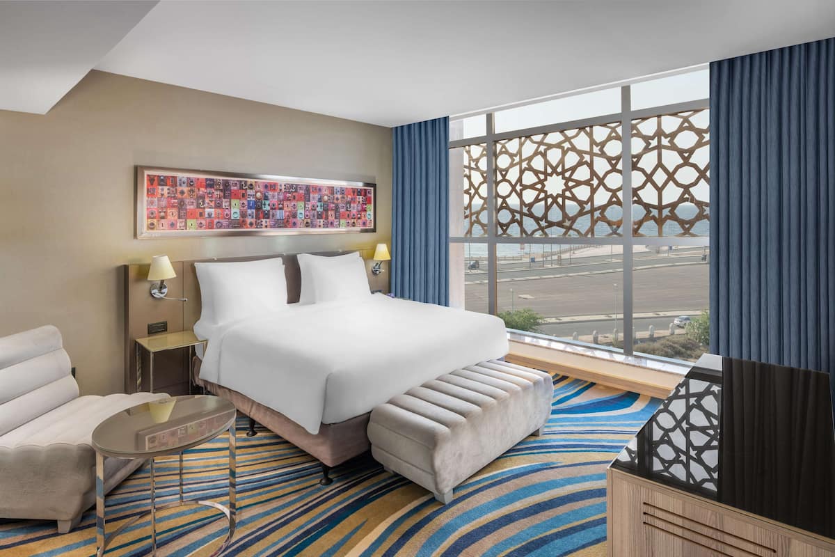 A hotel room with a large bed, a sofa, a small table, and a view of the city through a large window with a decorative screen.