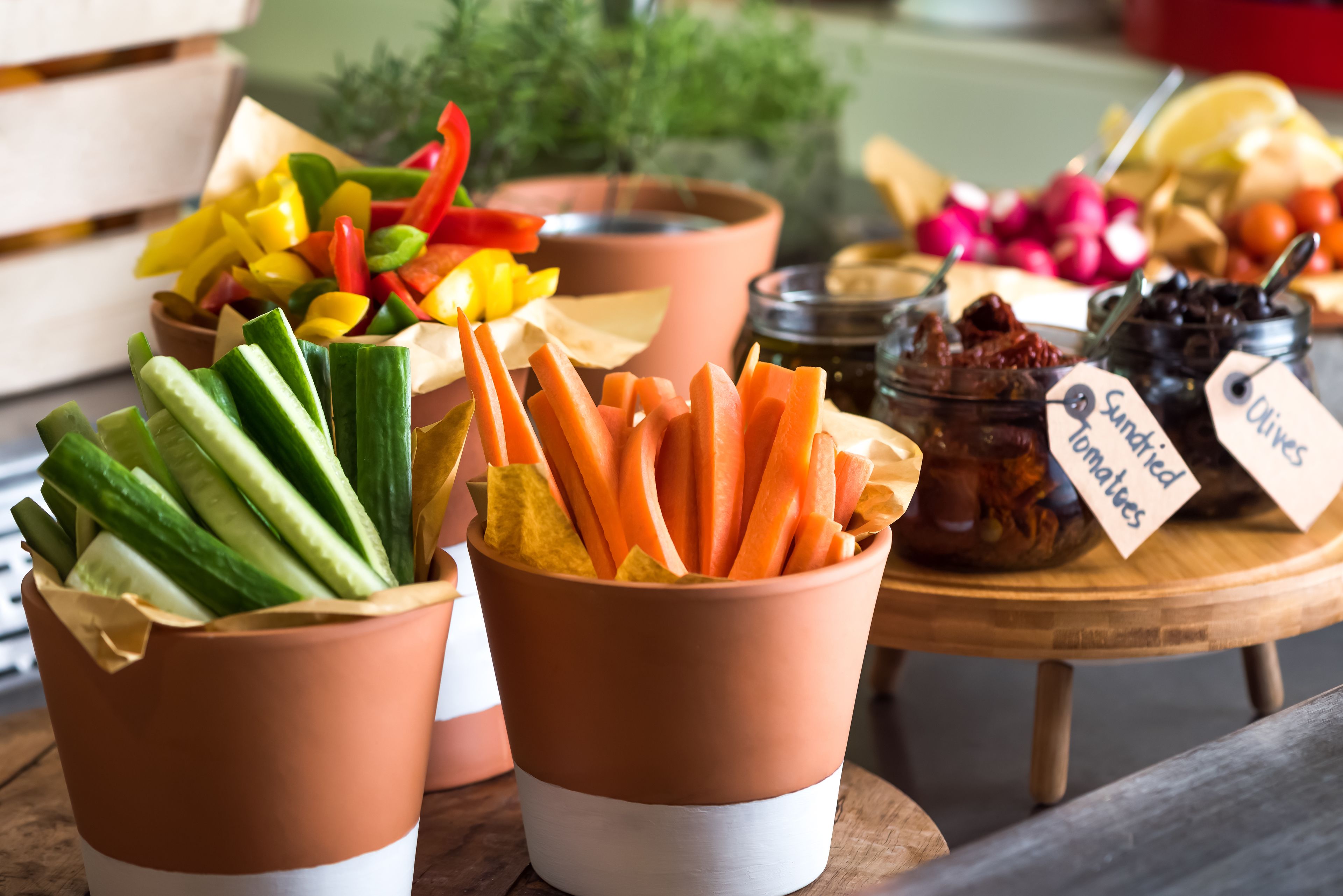 A variety of fresh vegetables in terracotta cups, including cucumbers, carrots, and bell peppers, with labels indicating they are sun-dried tomatoes and olives.