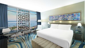 Une chambre d’hôtel avec un grand lit, un bureau avec une chaise, une vue sur l’océan par la fenêtre et un élément décoratif mural.