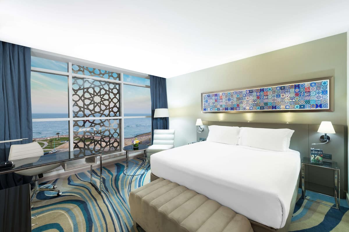 A hotel room with a large bed, a desk with a chair, a view of the ocean through a window, and a decorative wall piece.