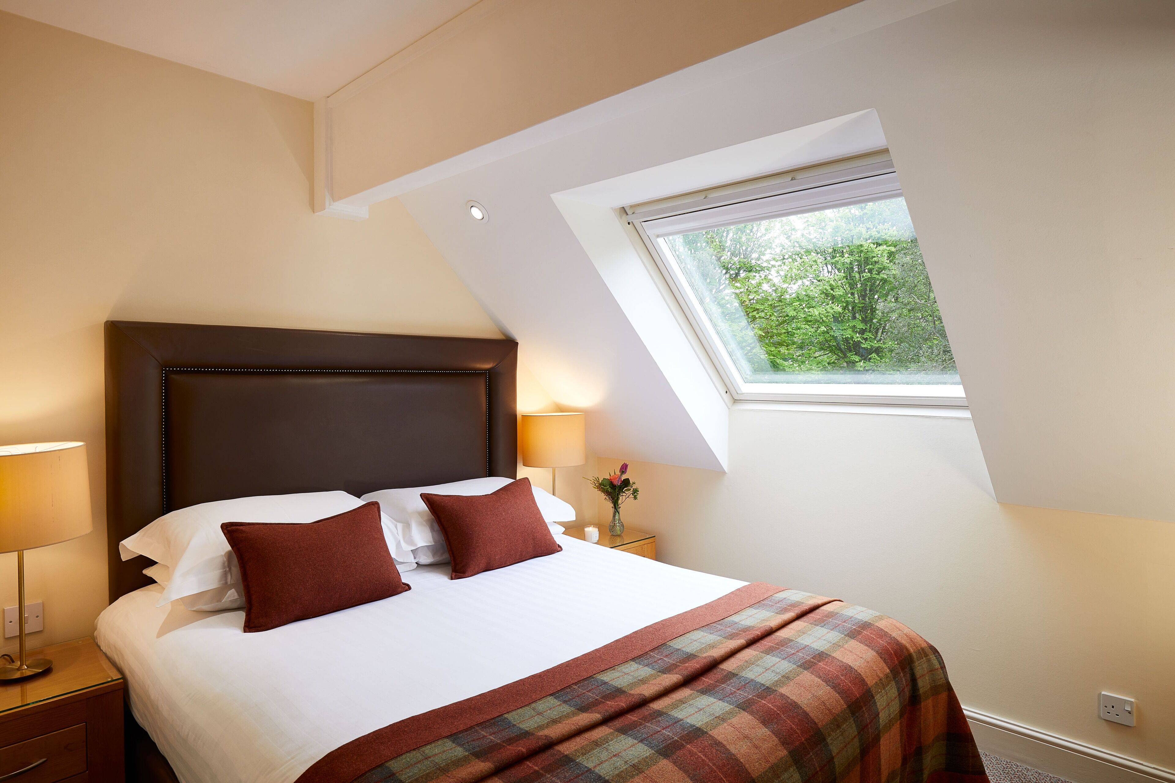 A hotel room with a large bed, a nightstand, a lamp, a window with a view of greenery, and a sloped ceiling.
