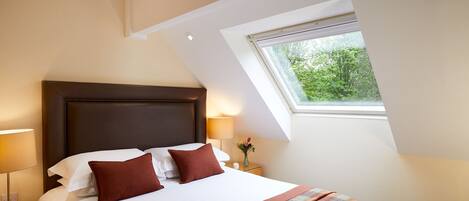 A hotel room with a large bed, a nightstand, a lamp, a window with a view of greenery, and a sloped ceiling.