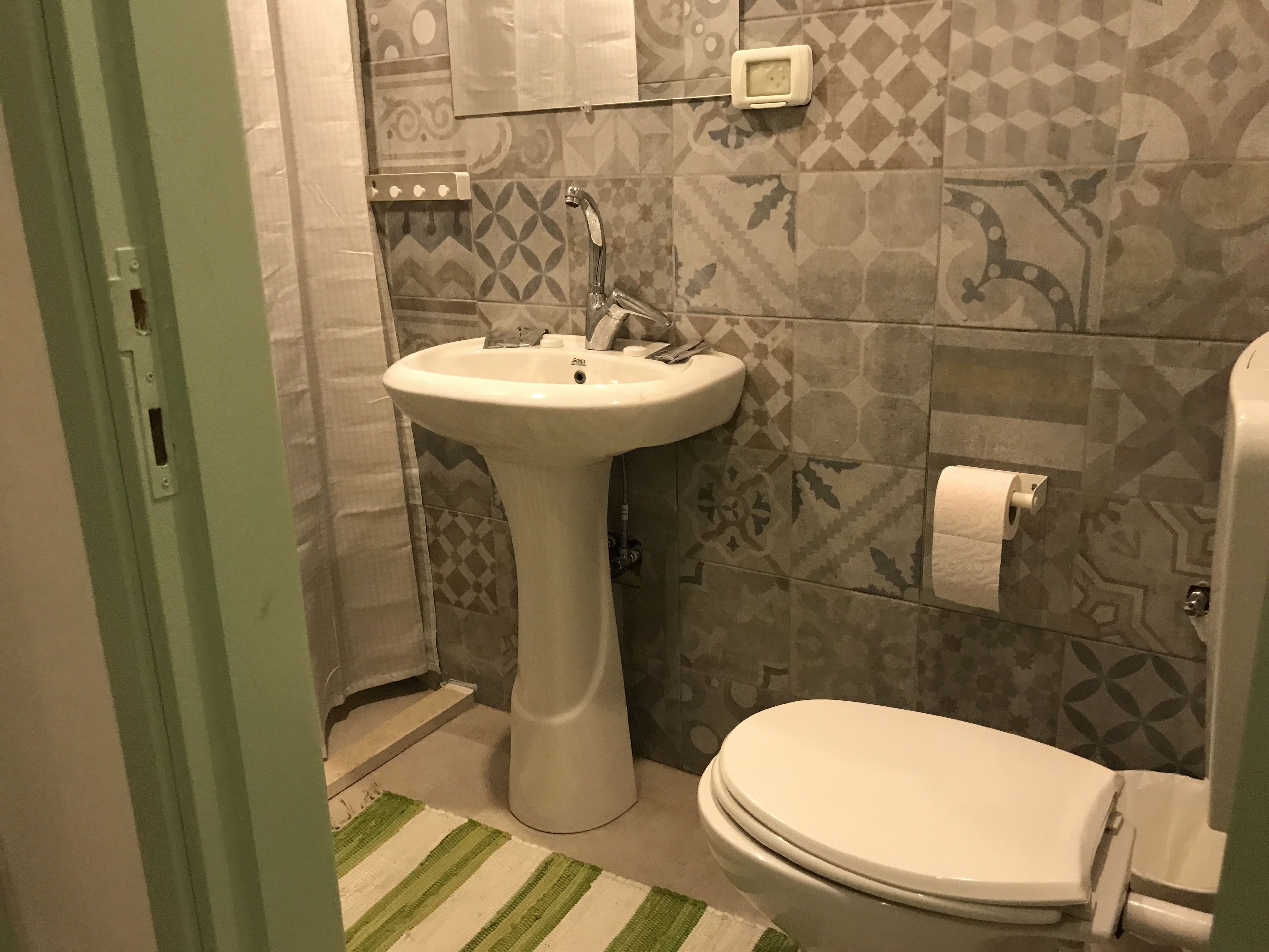 Un baño con lavamanos pedestal, inodoro, baldosas estampadas y un tapete verde a rayas.
