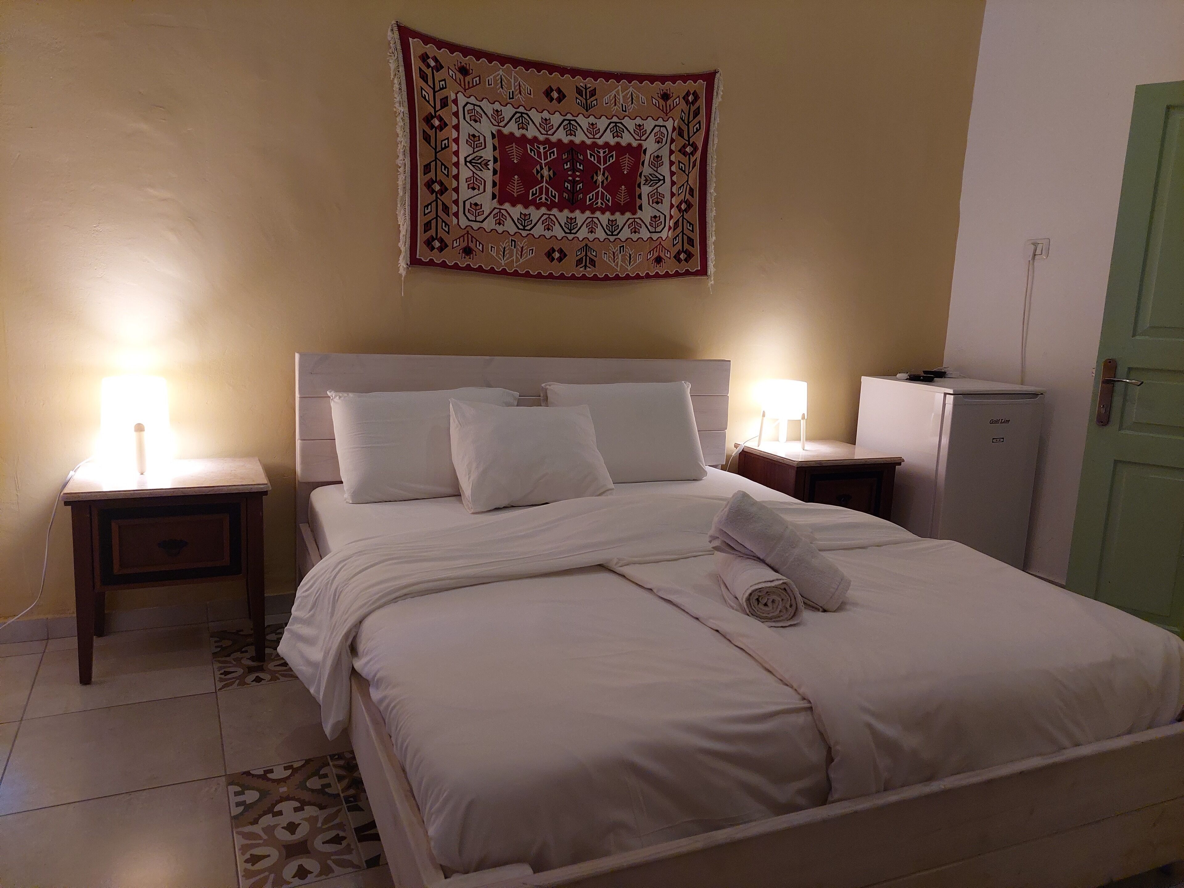 A neatly made bed with white linens, two bedside lamps, a small bedside table, a refrigerator, and a tapestry on the wall.