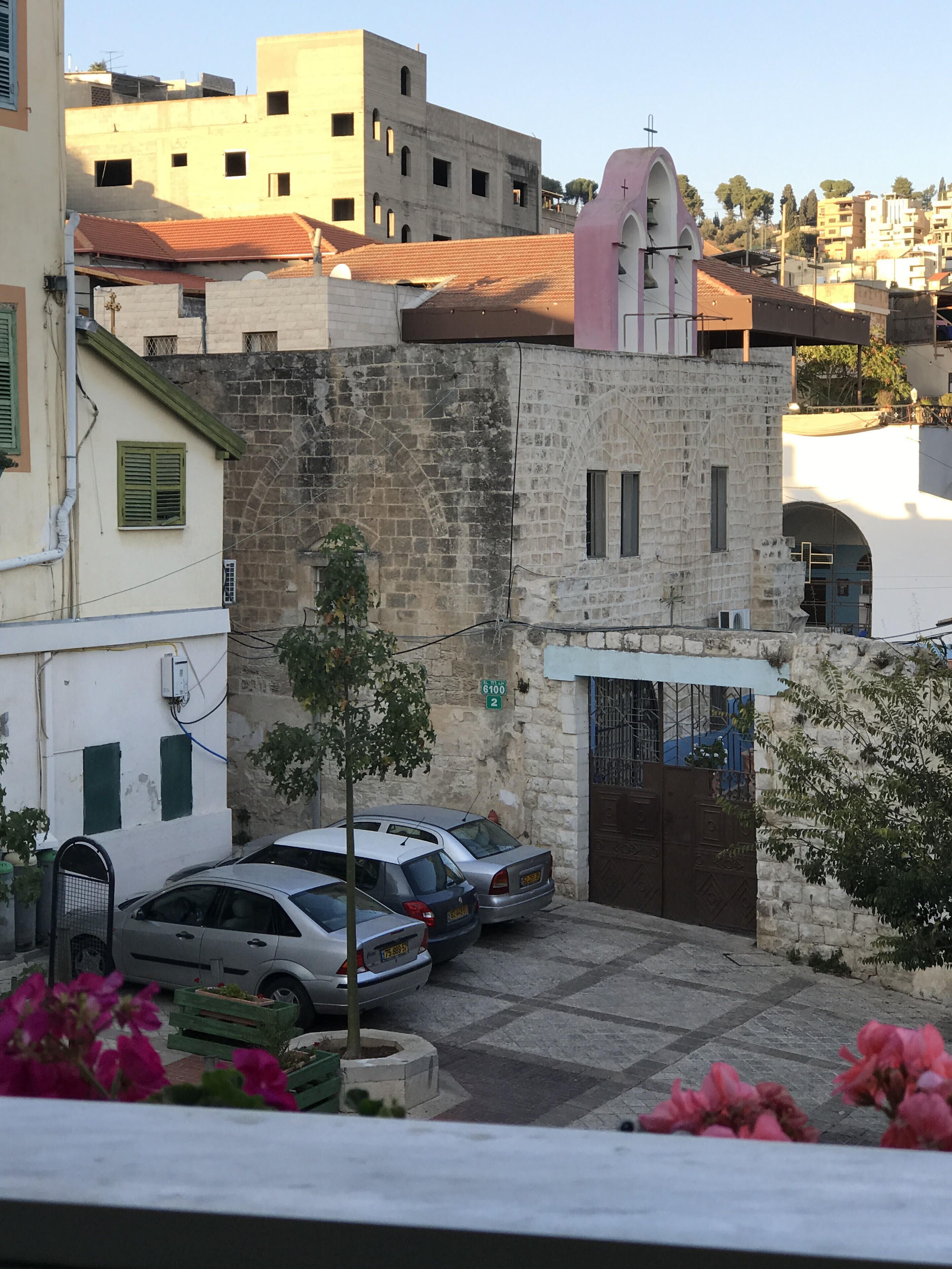 A street view with parked cars, a stone building with a gate, and a pink structure with a cross on top.