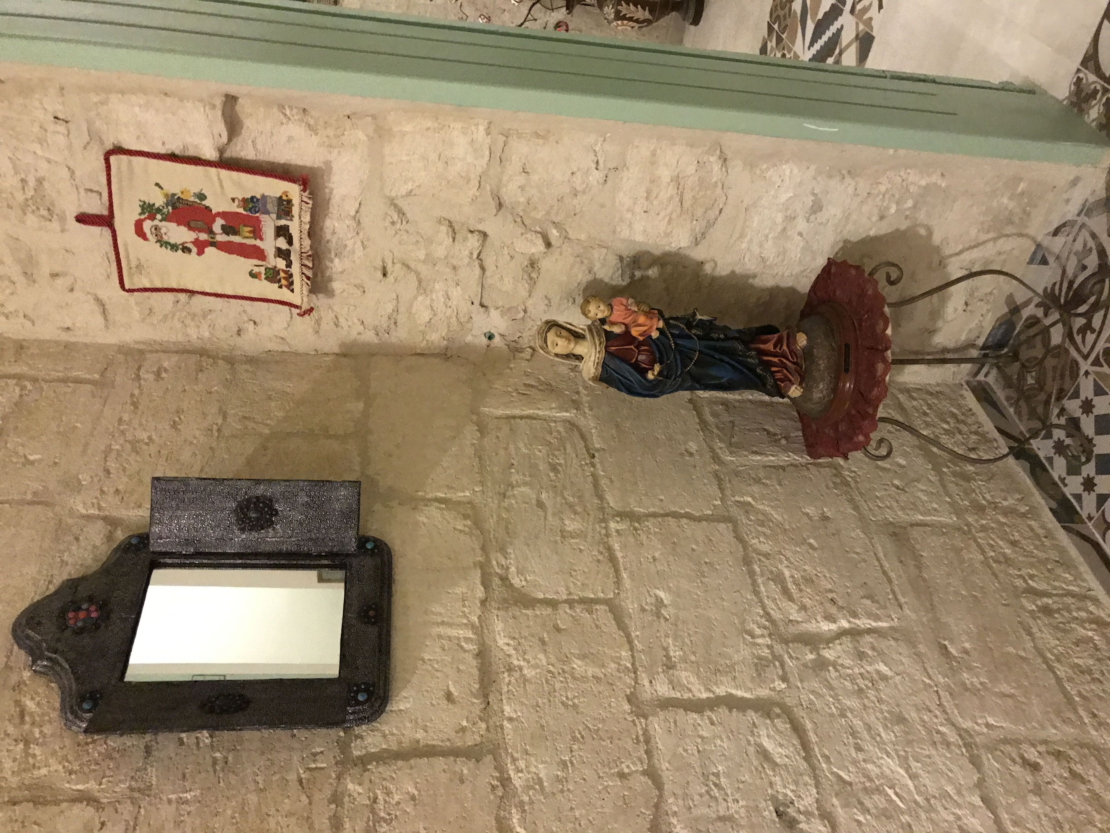 Un muro de piedra con una pequeña estatua de figura religiosa, un azulejo decorativo y un objeto negro que se asemeja a un control de juego.
