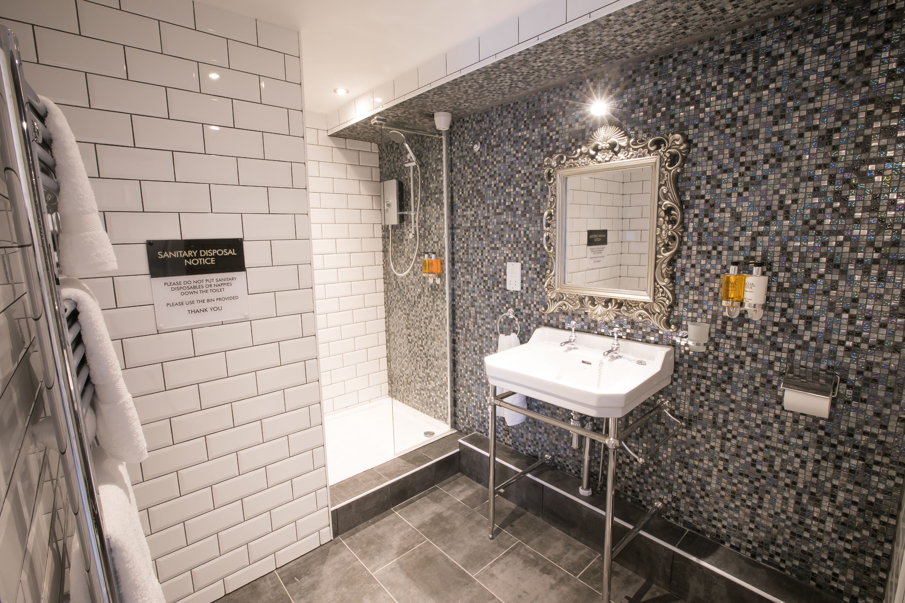 Une salle de bain moderne avec une douche, un lavabo et un miroir. Un panneau est affiché sur le mur, indiquant qu’il est interdit de jeter les articles d’hygiène féminine dans les toilettes.