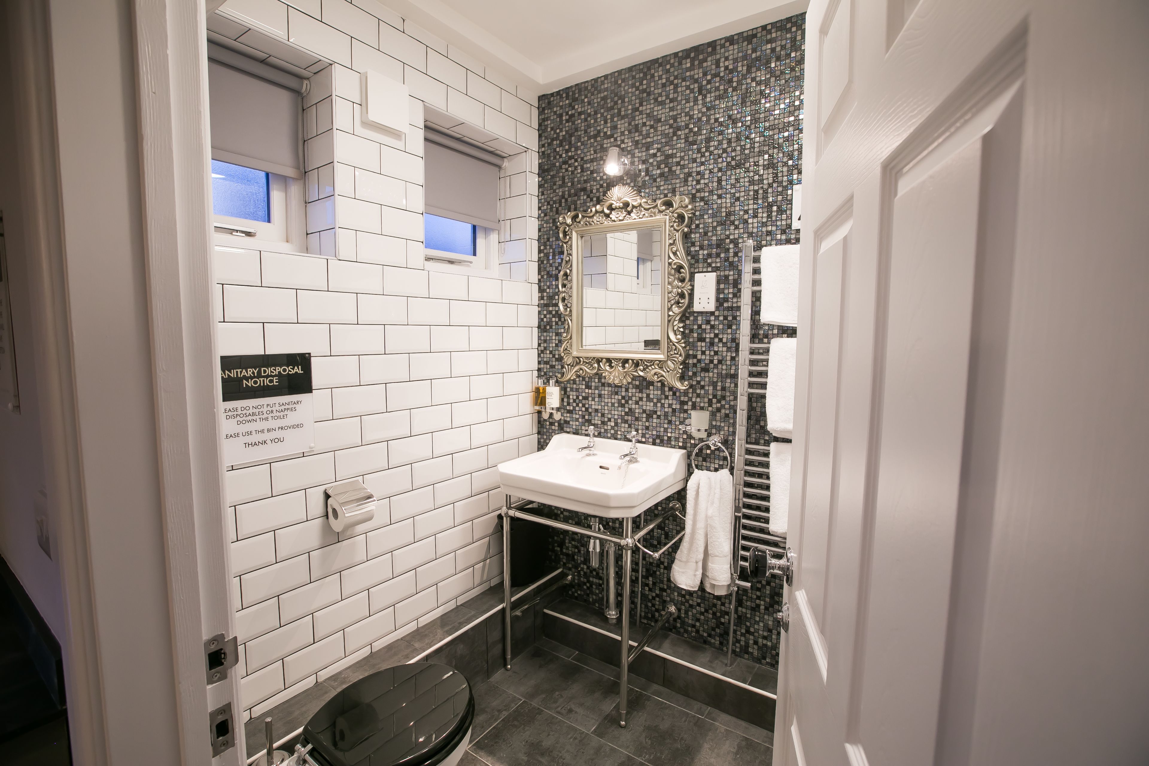 Un baño con paredes de azulejos blancos, un salpicadero negro de azulejos, un lavamanos blanco con espejo y un inodoro.