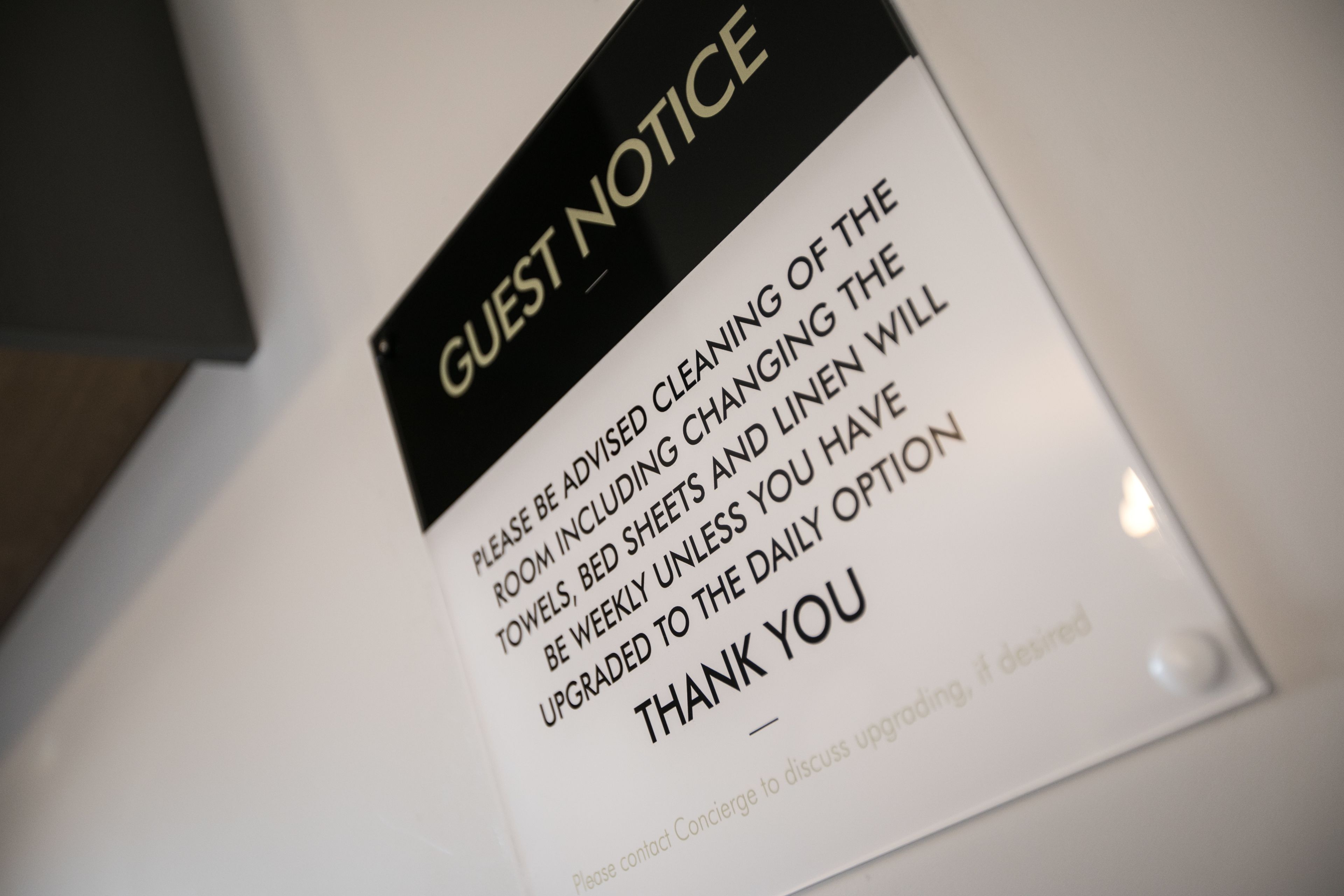A sign with a black top and white background. It reads "GUEST NOTICE" in bold letters. Below, it states "PLEASE BE ADVISED CLEANING OF THE ROOM INCLUDING CHANGING THE TOWELS, BED SHEETS AND LINEN WILL BE WEEKLY UNLESS YOU HAVE UPGRADED TO THE DAILY OPTION" followed by "THANK YOU".
