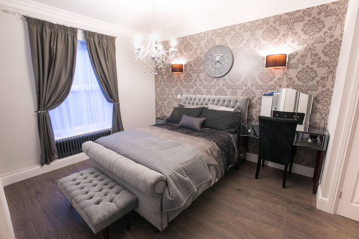 A bedroom with a large bed, a bedside table, a chair, a bench, a chandelier, and a clock on the wall.