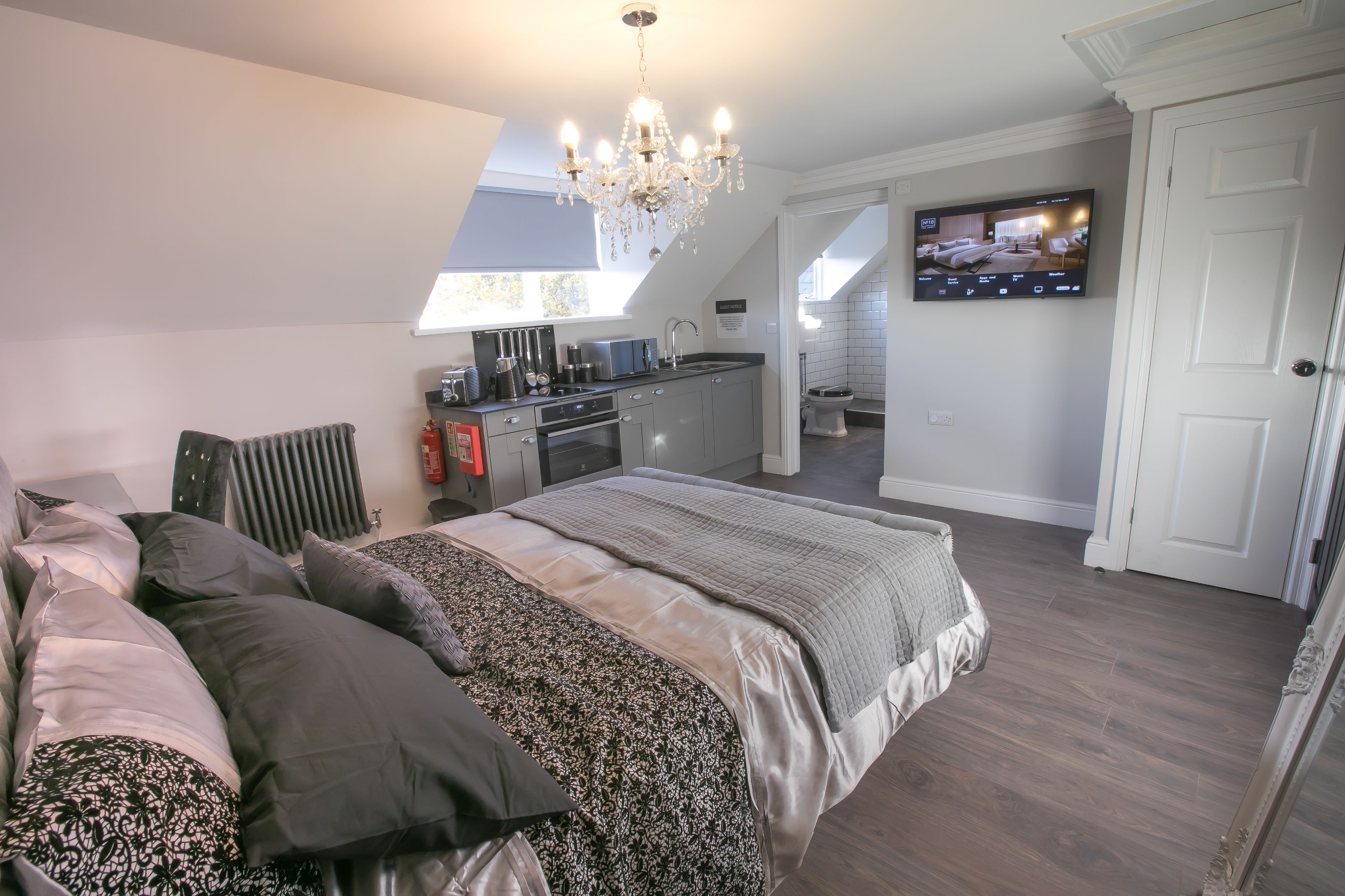 A bedroom with a large bed, a chandelier, a TV, and a kitchenette.