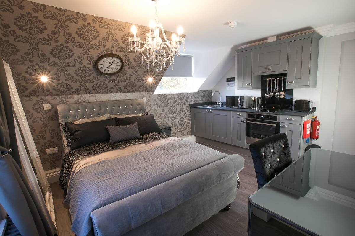 A bedroom with a large bed, a chandelier, a clock, and a view of the kitchen.