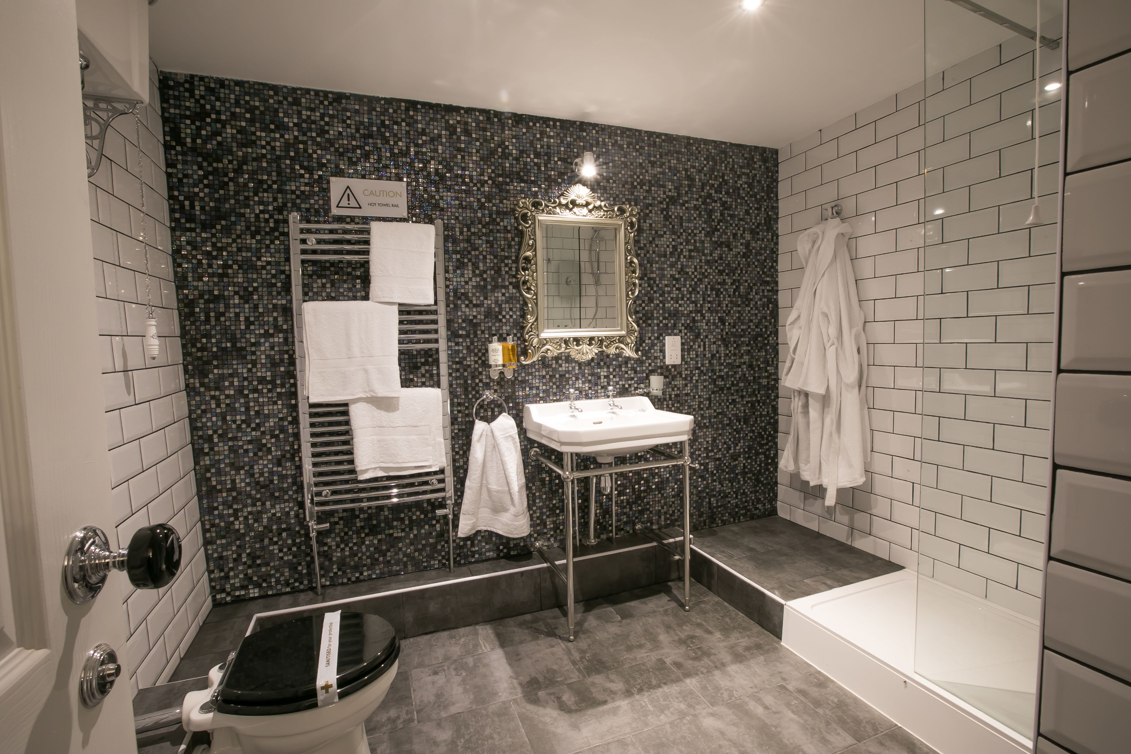 Et badeværelse med væg i sort mosaik, et hvidt håndvaskarrangement, et spejl, en håndklædestang og et toilet.